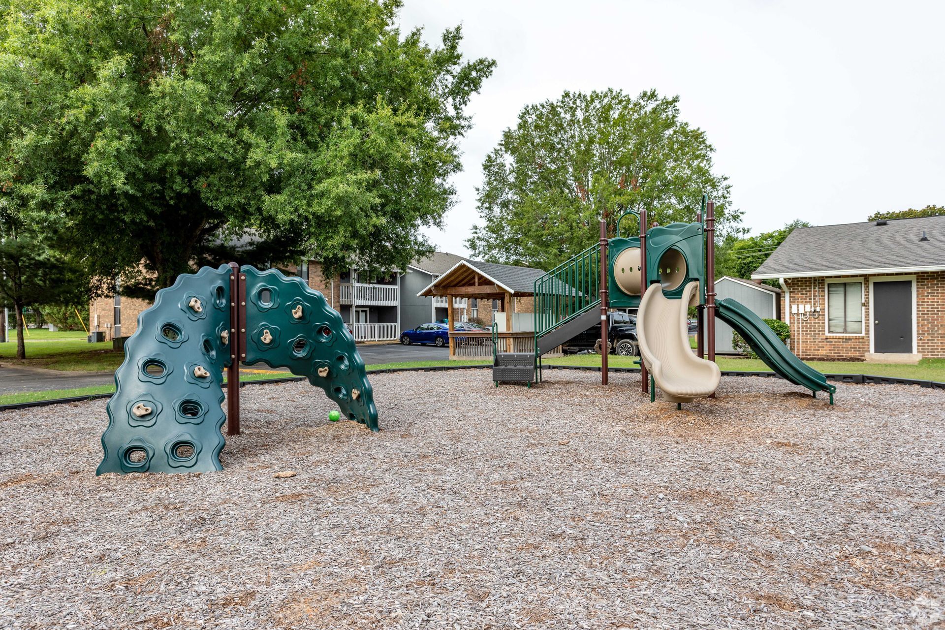 A playground with a green climbing structure, a central play set with a slide, and a wooden gazebo on wood chip ground.