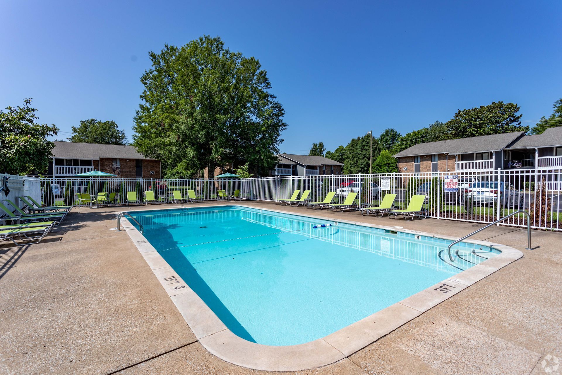An outdoor swimming pool surrounded by a concrete deck, lounge chairs, a white fence, and apartment buildings.