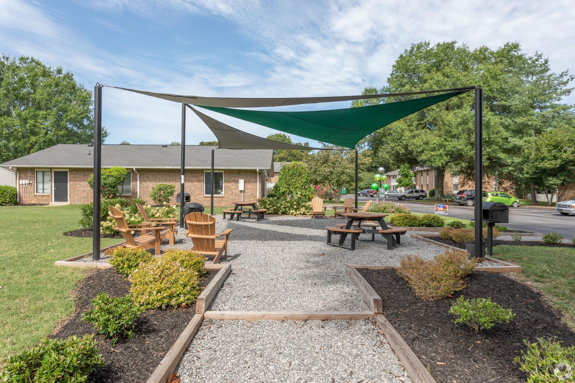 A gravel courtyard with wooden Adirondack chairs and picnic tables under a green and beige triangular sun shade.
