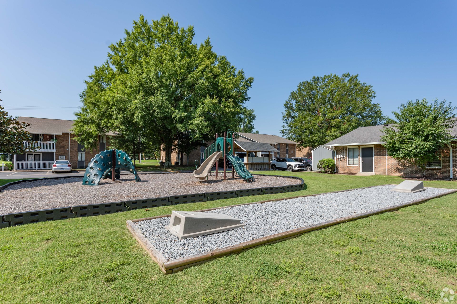 A playground with a climbing structure and slide set in gravel, with an adjacent narrow gravel pit and apartment buildings.