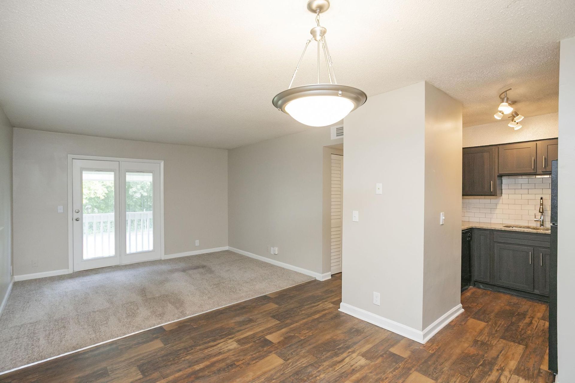 A dining and living area with grey carpeting, wood-style flooring, a modern pendant light, and a glimpse of a kitchen.