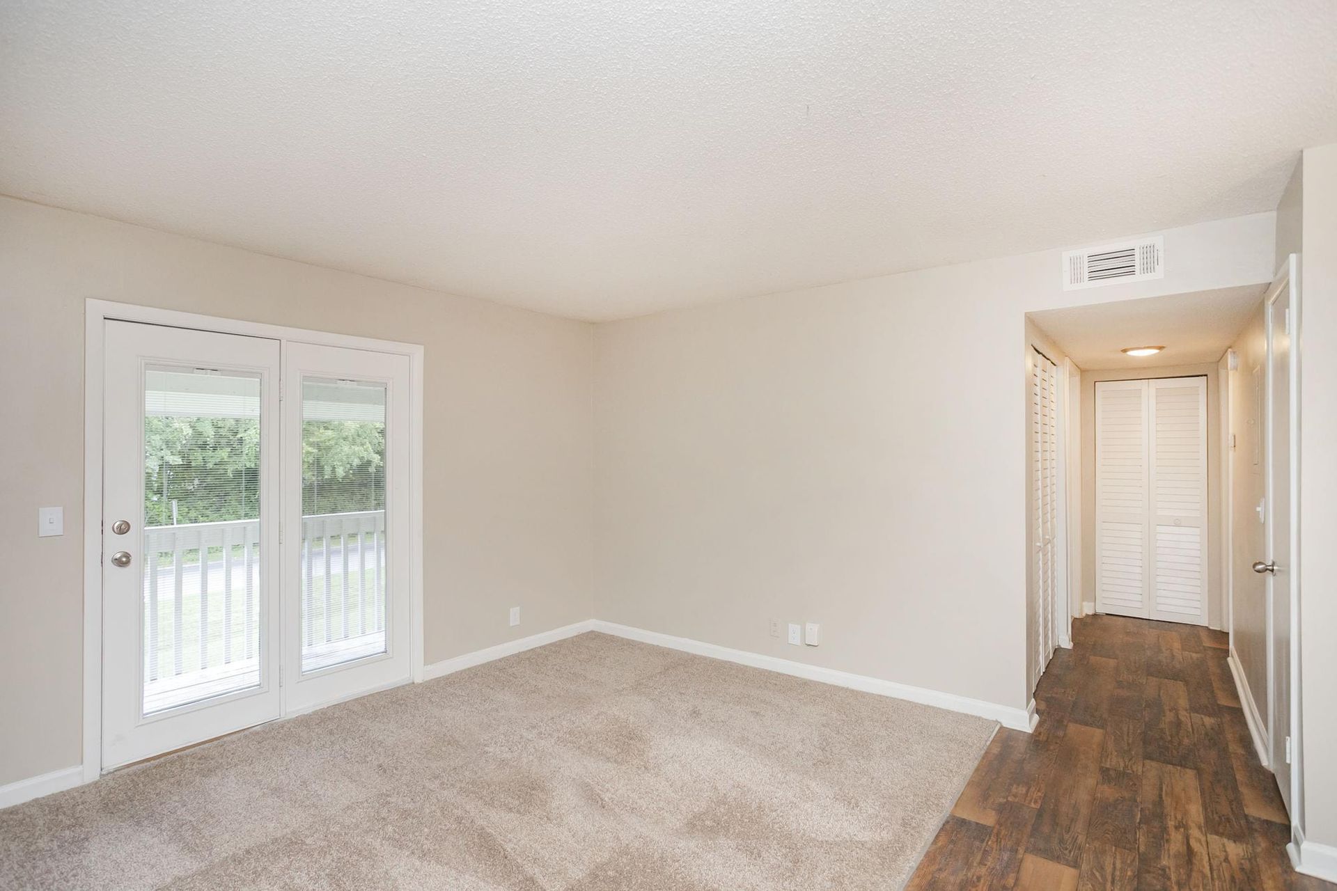 A beige room with carpet, a glass door leading to a balcony, and a hallway transition to a wood-floored area.