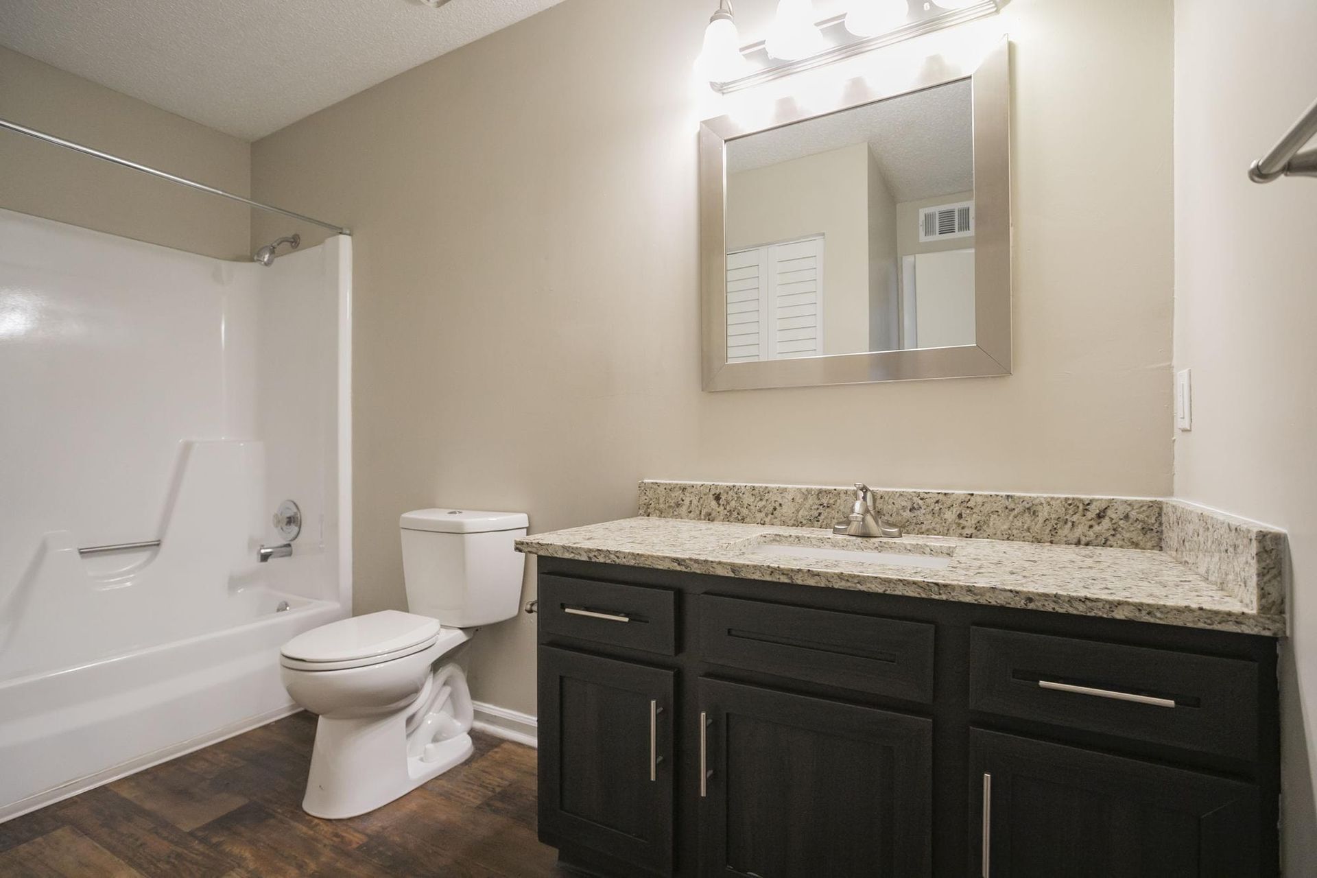 A bathroom with a dark wood vanity, granite countertop, white toilet, and a white shower-tub unit against beige walls.