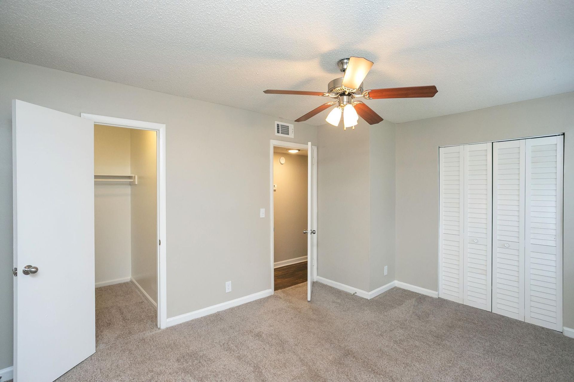 A beige room with grey carpet, a ceiling fan, an open closet, an open door to a hallway, and louvered closet doors.