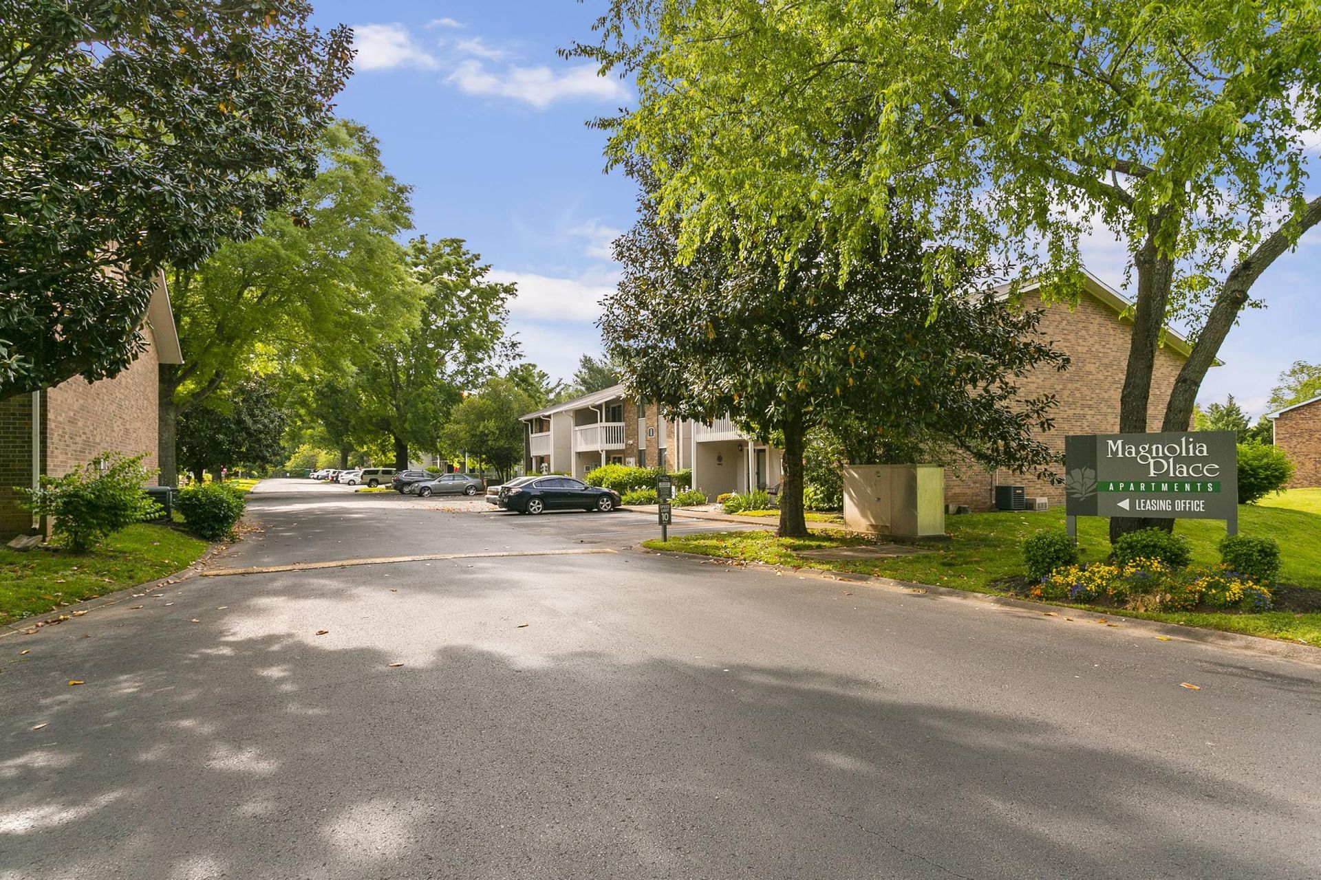 An apartment complex driveway with mature trees, green lawns, and a wooden entrance sign under a sunny blue sky.