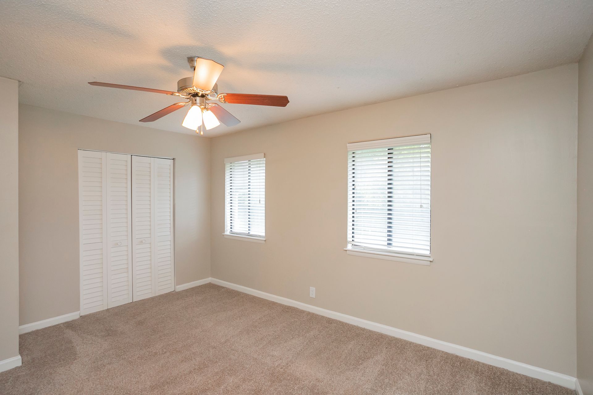 An empty bedroom with beige walls, brown carpet, a ceiling fan, and white louvered closet doors under two windows.