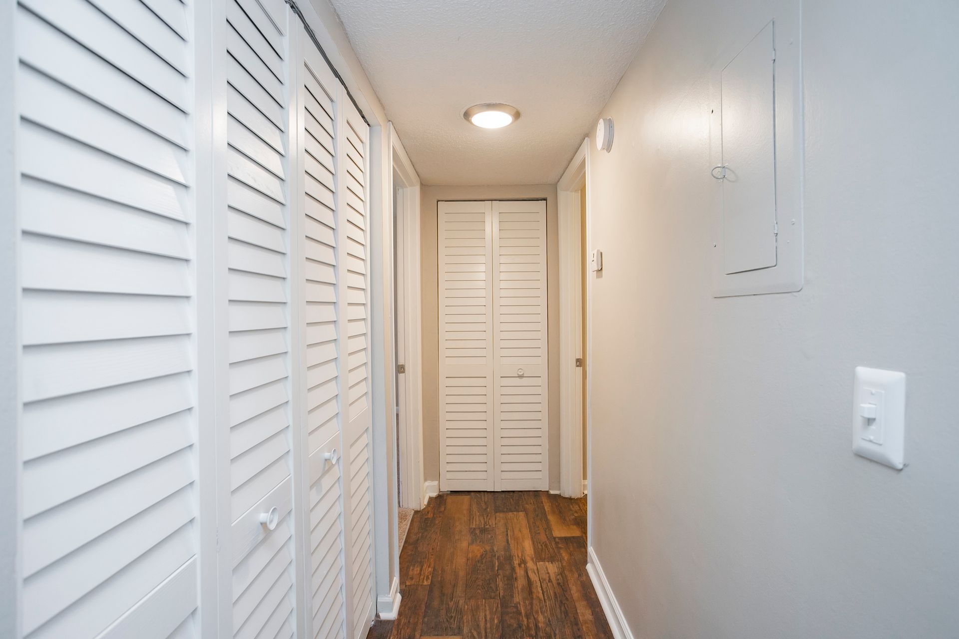 A hallway featuring white louvered doors on the left, a light switch on the right, and a door at the end of the passage.