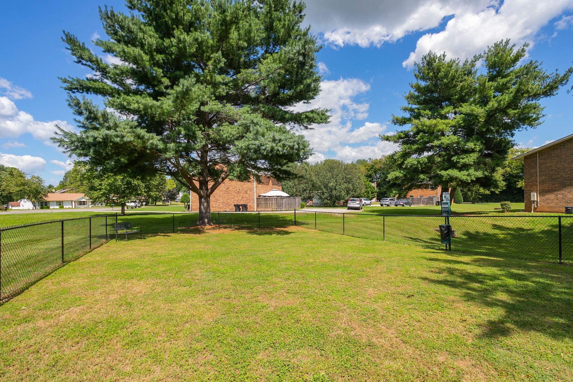 A grassy fenced dog park area with two large trees and a brick building under a blue sky with fluffy white clouds.
