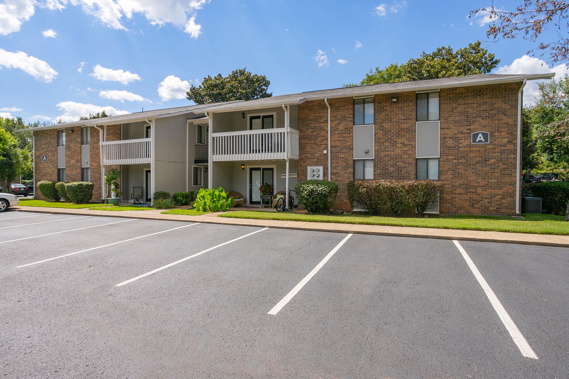 A two-story brick apartment building with white balconies sits behind an empty asphalt parking lot under a blue sky.