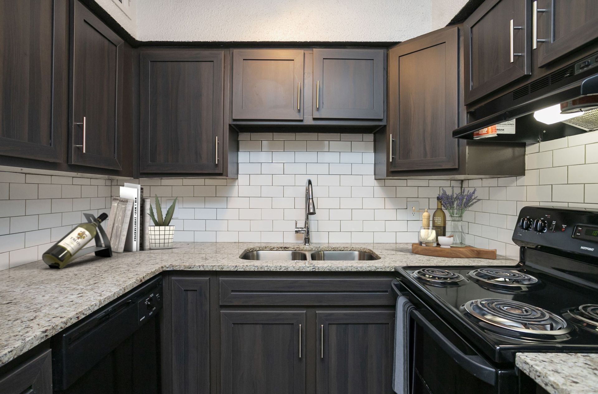 A kitchen with dark wood cabinets, light granite countertops, white subway tile backsplash, and black appliances.