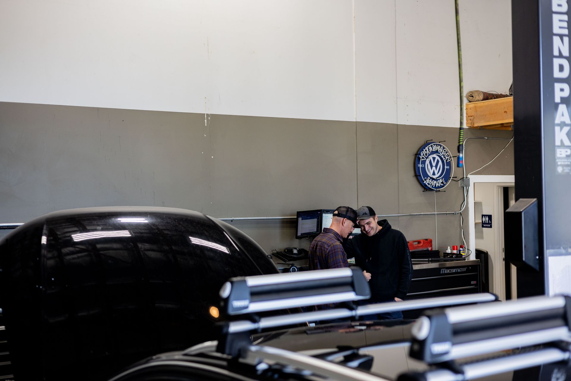 Two men by a car inside a garage; one pointing at something. Silver ski racks on the black car roof.| Peak Euro