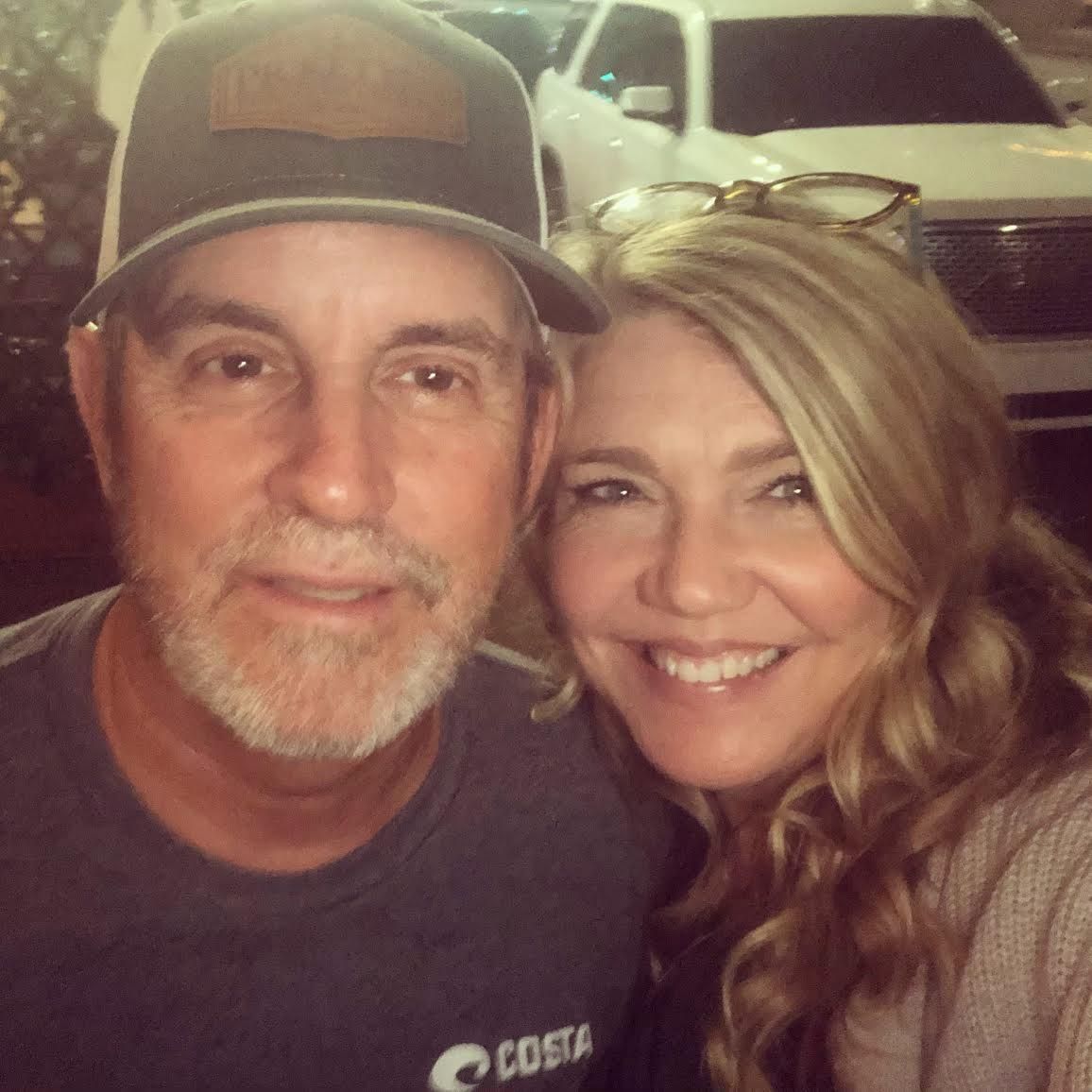 Man and woman smiling, close-up selfie. Man wears hat, gray shirt. Woman with blonde hair. Vehicle in background.