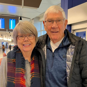 Couple in airport, smiling. Woman wears scarf, glasses. Man wears blue sweater, glasses, black jacket.