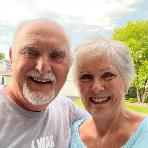 Smiling couple, outdoors. Man with white beard, wearing gray shirt. Woman with short white hair, blue top.