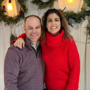 Couple smiling, woman in red turtleneck, man in purple sweater, festive background.