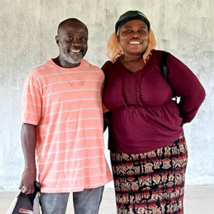 Two smiling people standing against a plain wall, one in a pink striped shirt and one in a maroon top and patterned skirt.