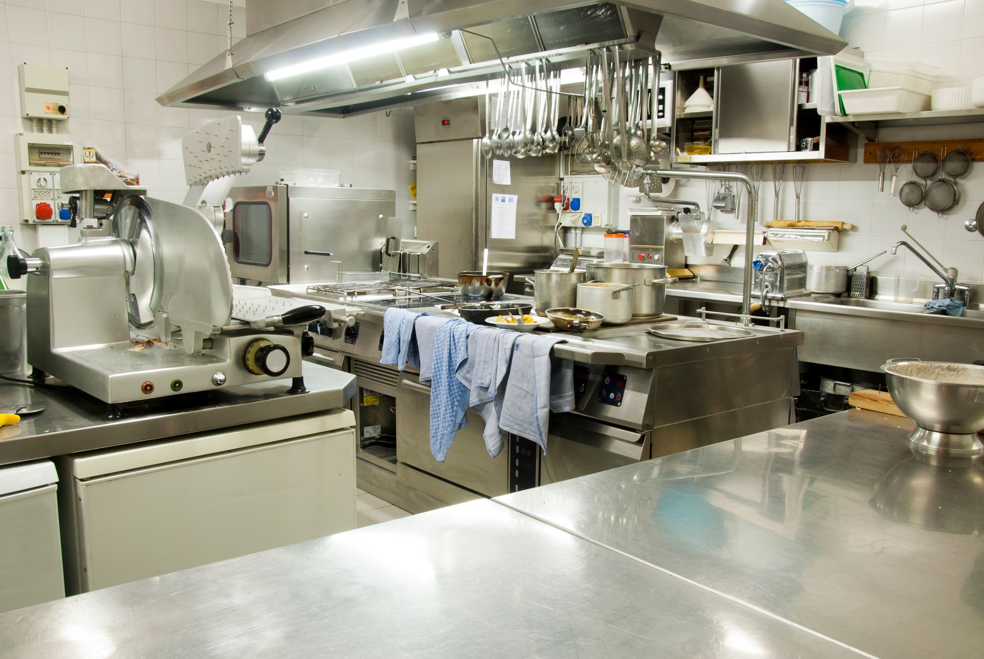 Commercial kitchen with stainless steel counters, pots, utensils, and hanging towels under bright lights.