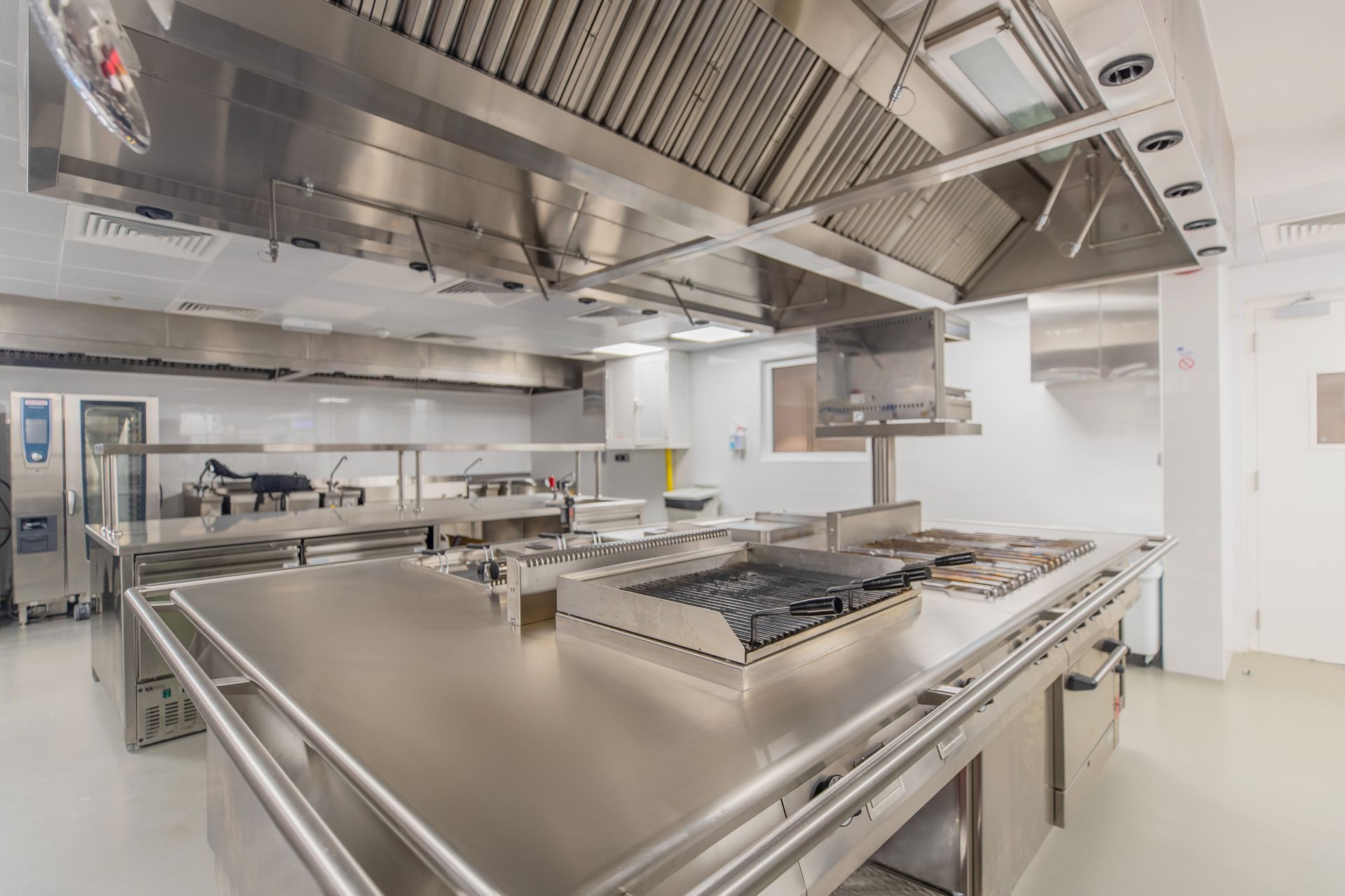 Modern commercial kitchen with stainless steel prep tables, stovetops, and ventilation hoods.