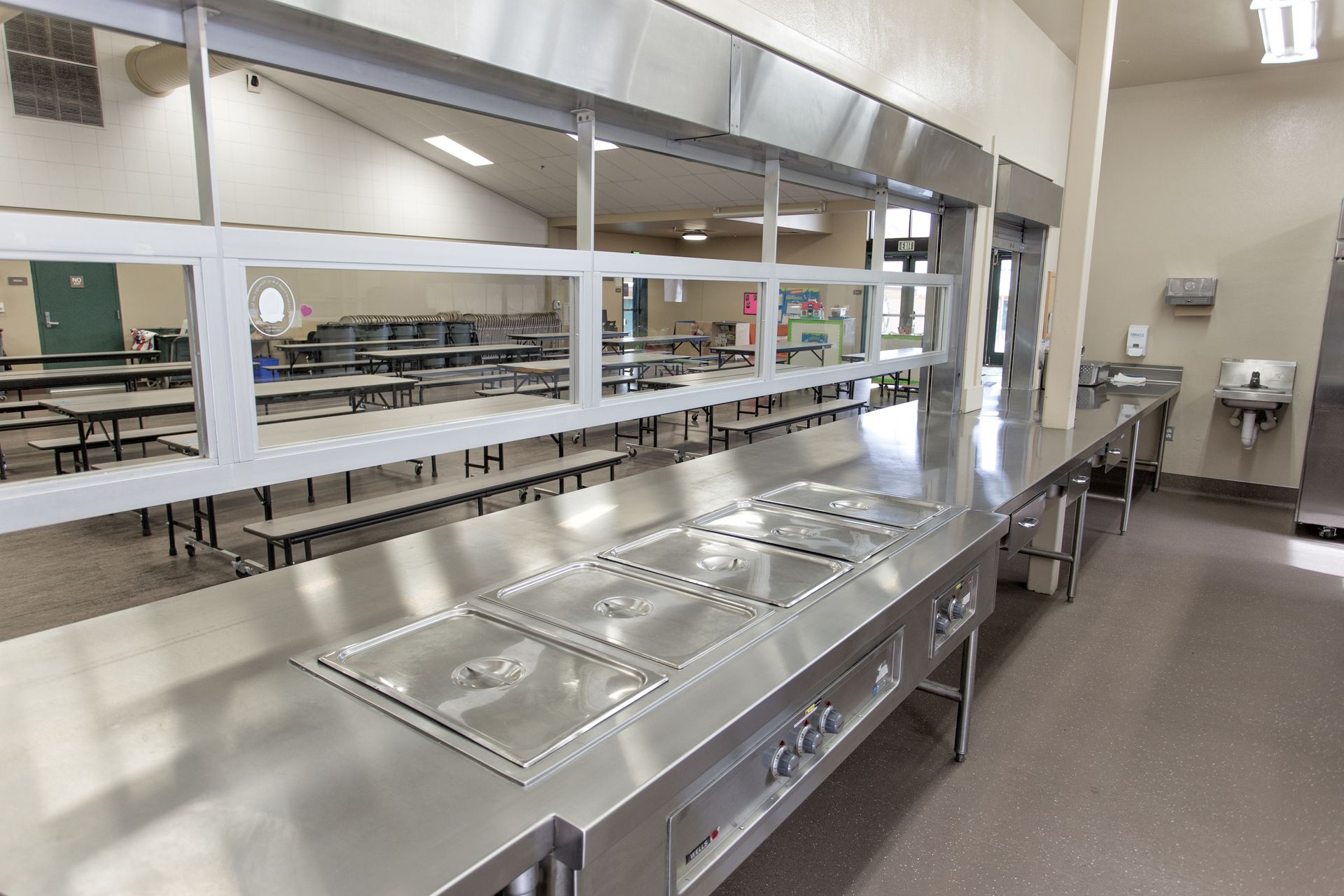 Empty school cafeteria with stainless steel serving counters and benches in a bright dining hall.