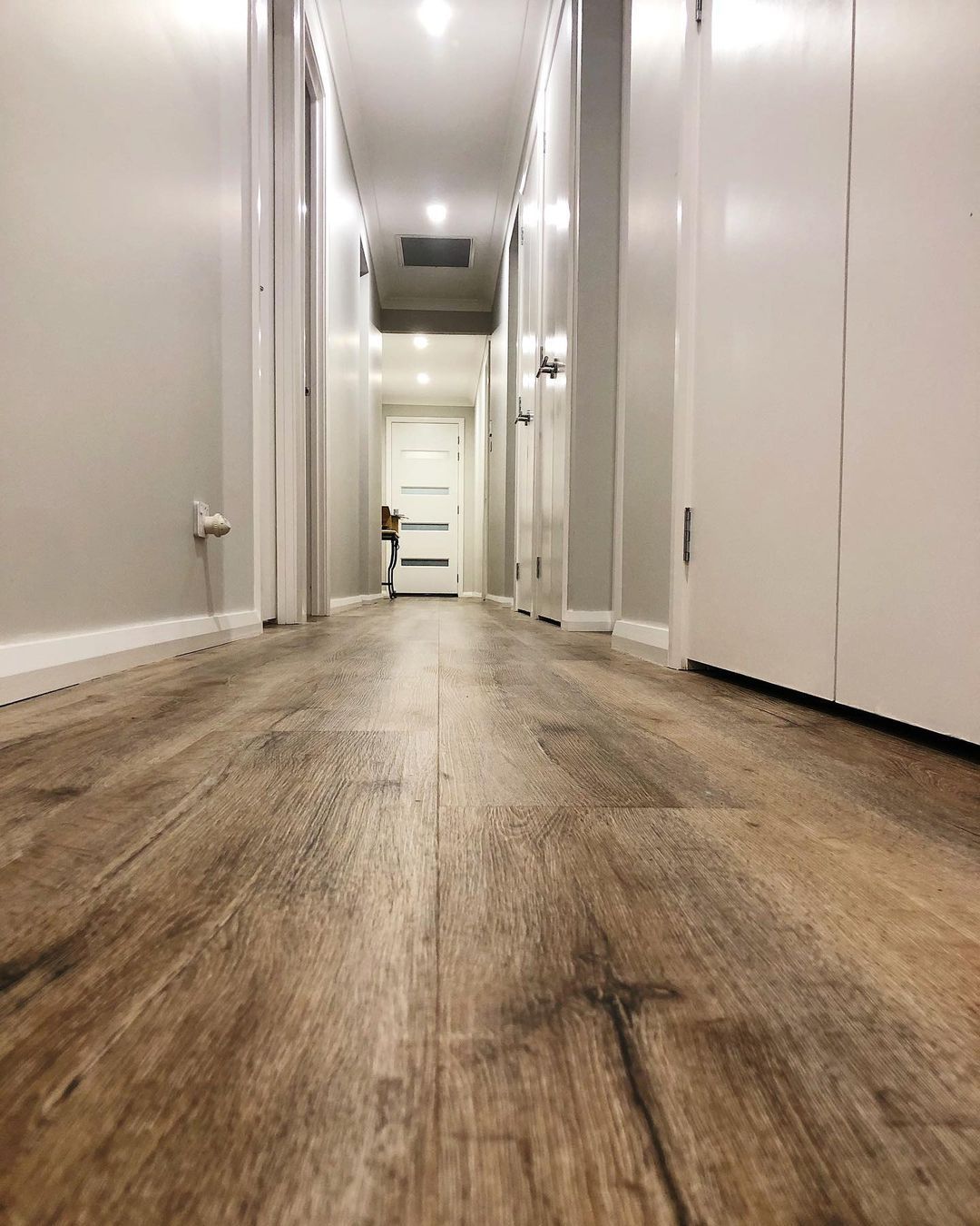Narrow hallway with wood-look floor, white walls, and doors leading to a bright room at the end.