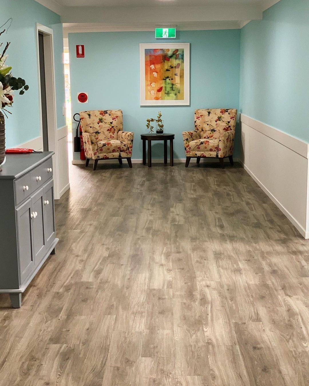 Quiet hallway with two floral armchairs, a small table, and light blue walls under an exit sign.