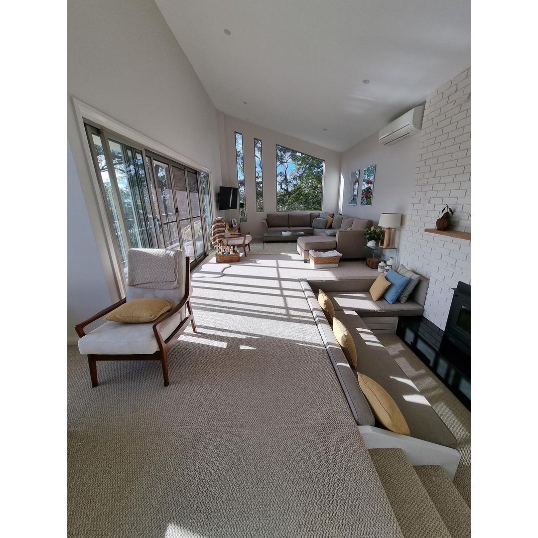 Bright sunroom with woven rug, white chairs, beige sofa, and large windows casting striped sunlight.