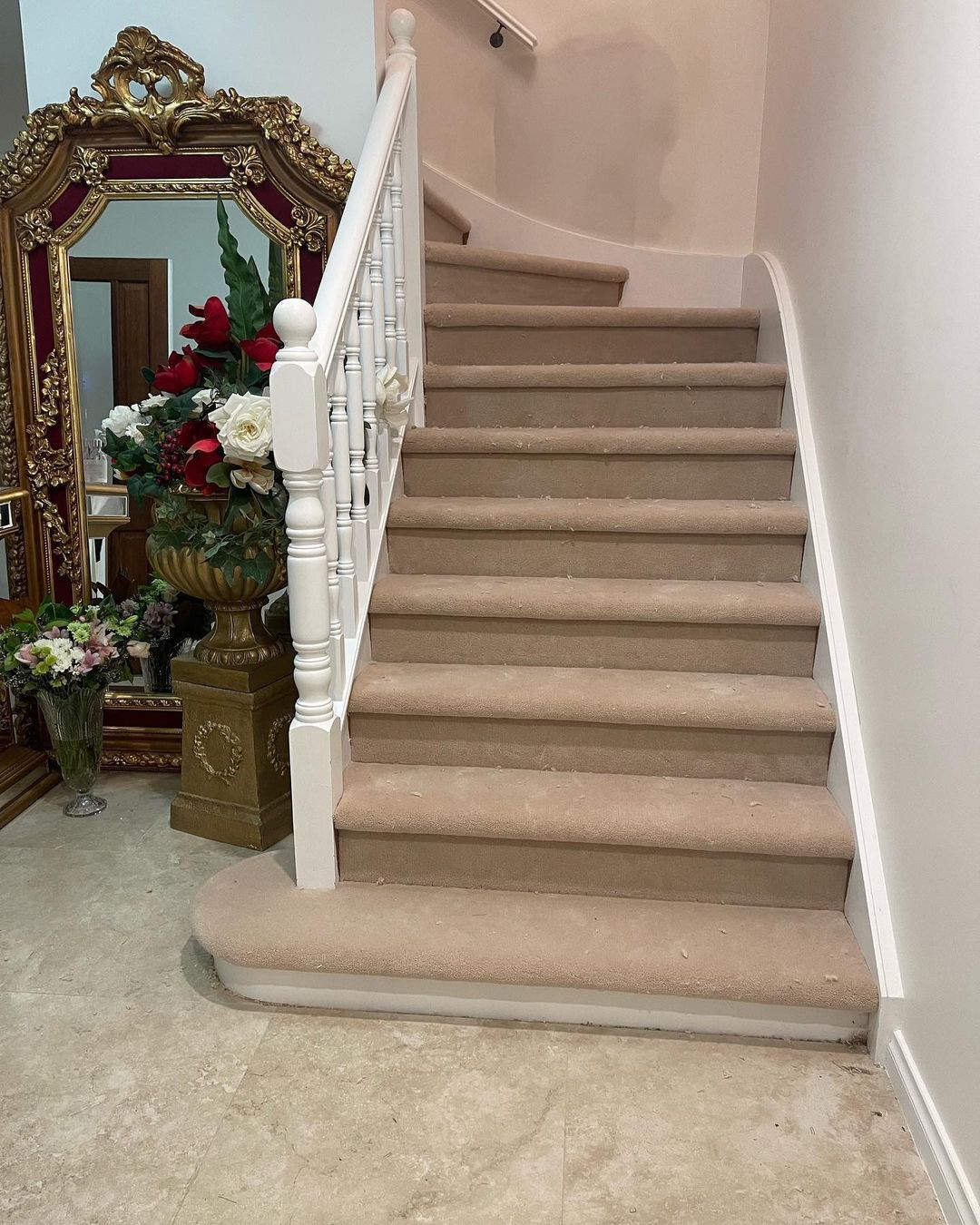Carpeted staircase with white railing in a beige home interior, beside a decorative mirror and floral arrangement