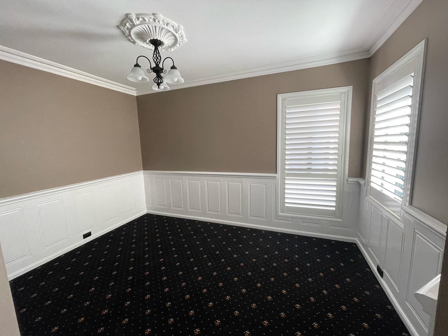 Empty dining room with beige walls, white wainscoting, black carpet, and two windows with white blinds