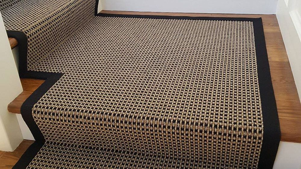 Black woven rug with geometric pattern covering stairs and landing in a hallway