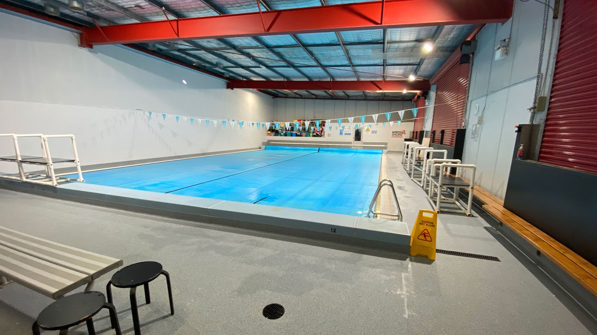 Indoor swimming pool with lane lines, red ceiling beams, and chairs along the deck.