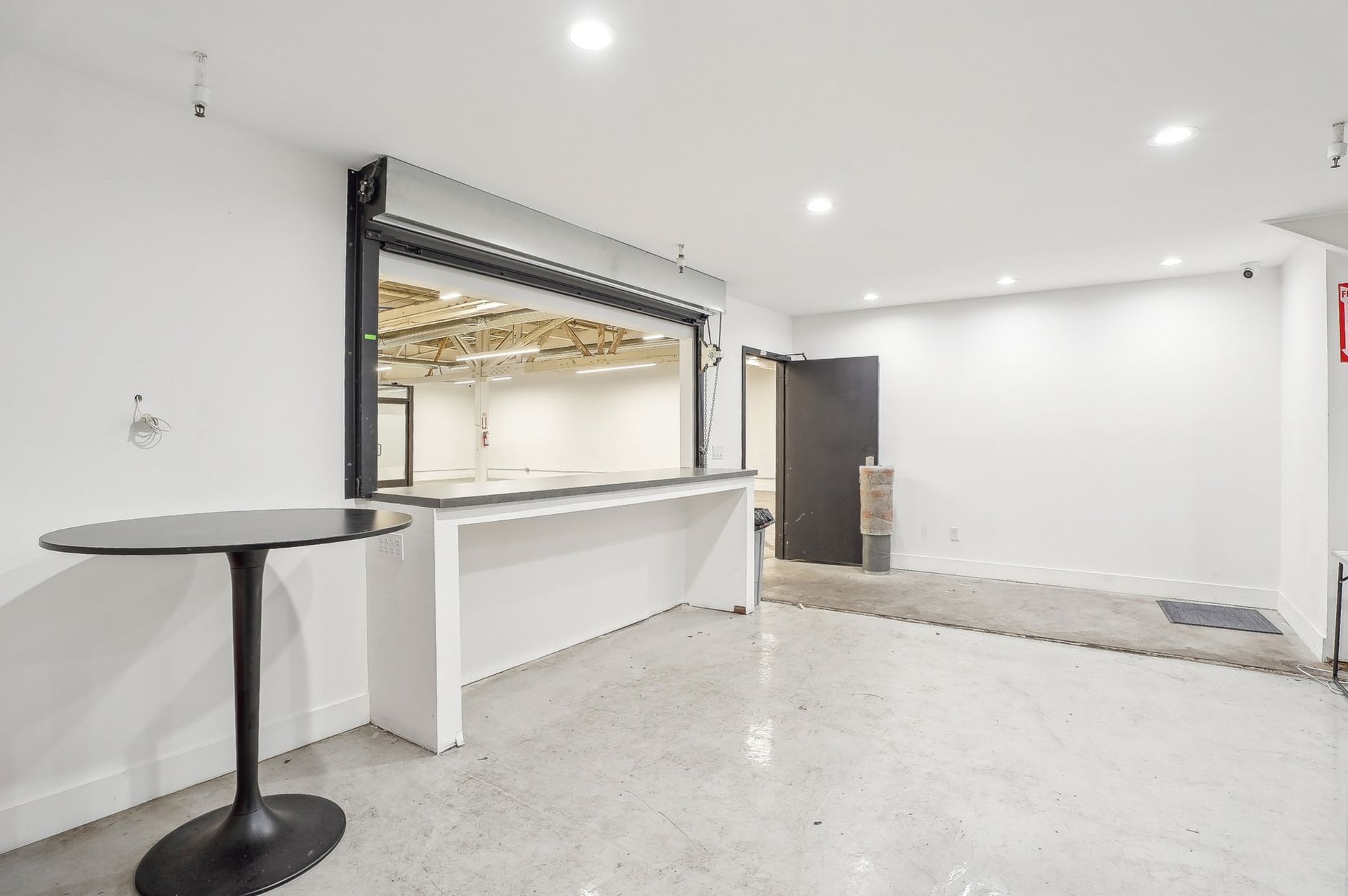 An empty white room with a service counter, a window looking into another room, and a small round table in the foreground.