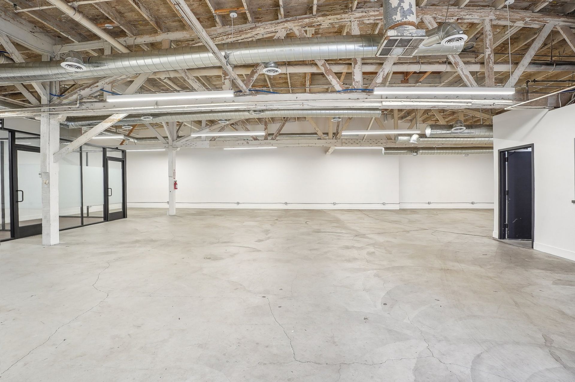 An empty, industrial-style commercial space with white walls, exposed ceiling beams, and concrete floors.