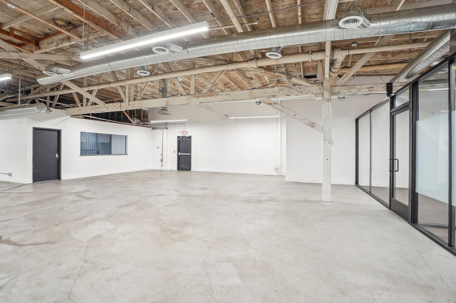 An empty industrial office space with concrete floors, exposed ceiling beams, white walls, and glass-partitioned rooms.