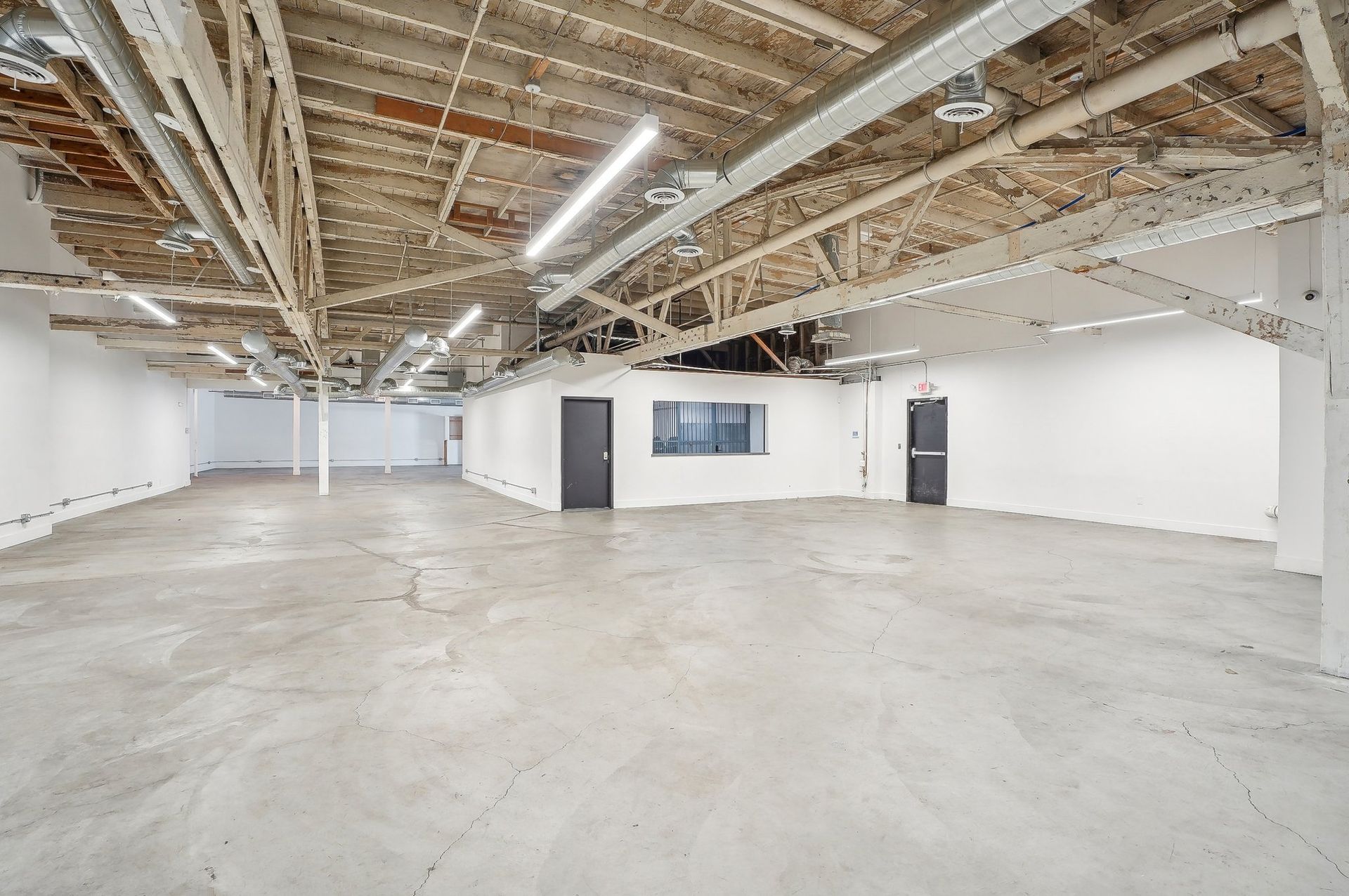 Wide, open industrial loft space with exposed wood ceiling beams, white walls, and a smooth, light-colored concrete floor.