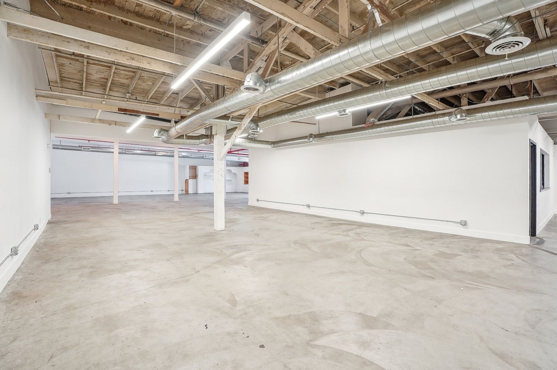 An empty industrial space with white walls, a concrete floor, exposed wooden ceiling beams, and exposed HVAC ductwork.