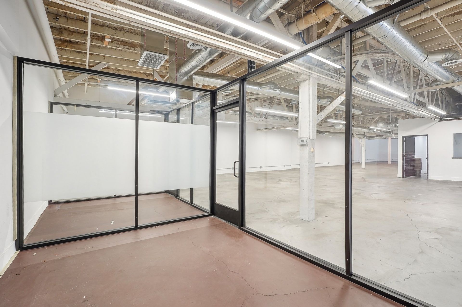 An interior view of a modern, open-plan office space with glass-walled partitions and exposed industrial ceiling pipes.