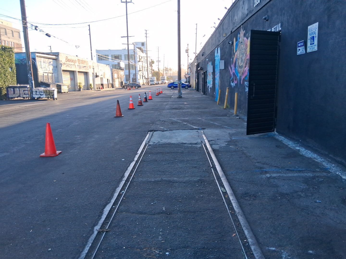 A narrow urban street with a row of orange traffic cones and a set of old metal tracks embedded in the pavement.