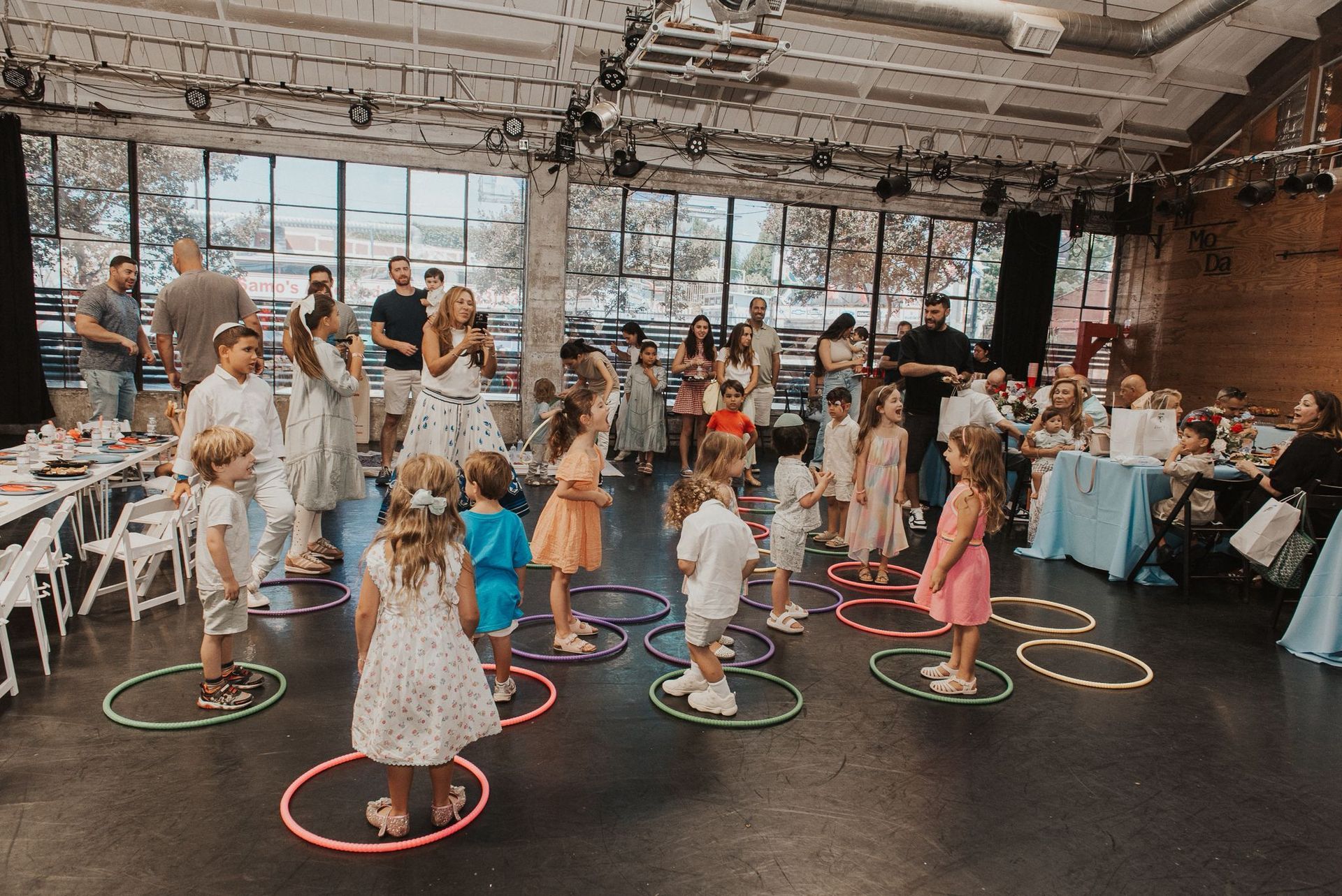 Children in a bright, spacious hall participate in a game, standing inside colorful hula hoops arranged on the floor.