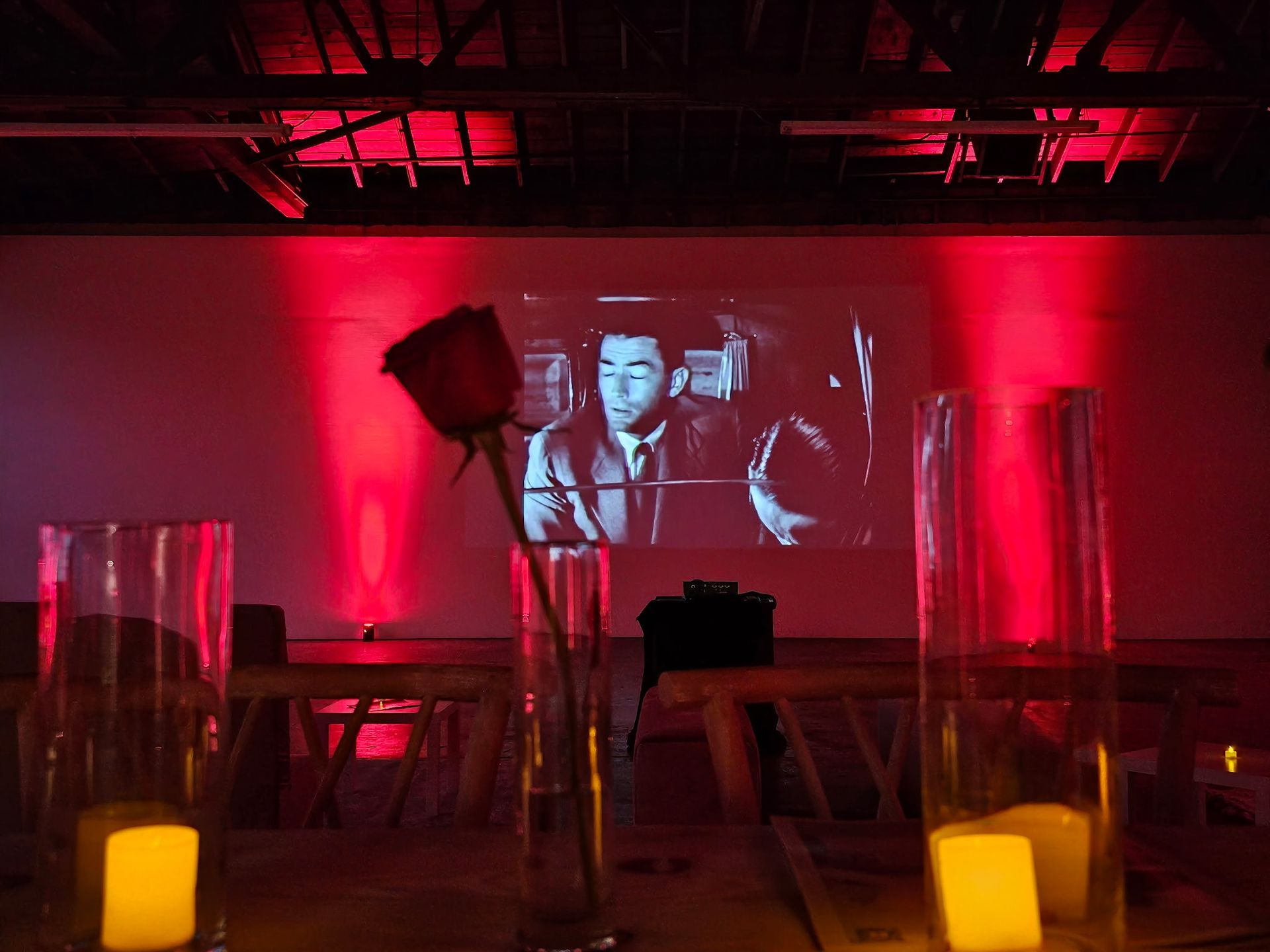 A dimly lit room with red mood lighting features a projected black-and-white film, a single rose, and glowing candles.