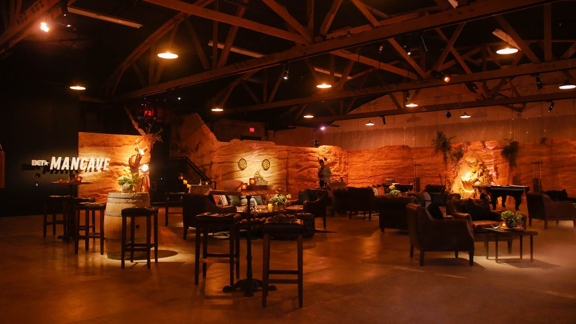 Dimly lit, industrial event space with dark furniture, exposed wooden ceiling beams, and warm, textured stone walls.