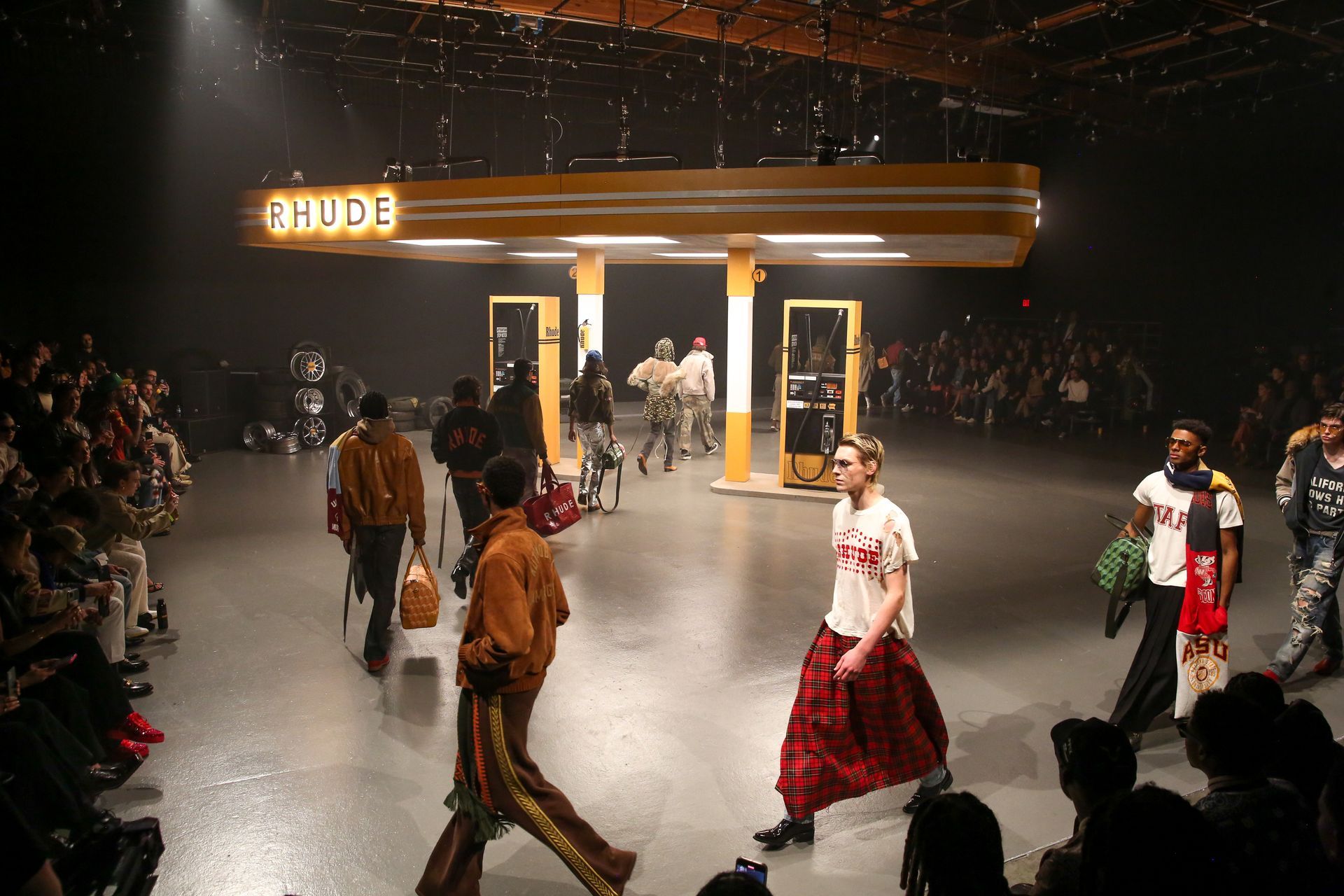 A fashion show features models walking on a runway set inside a dimly lit space with a large, neon-lit Rhude gas station.