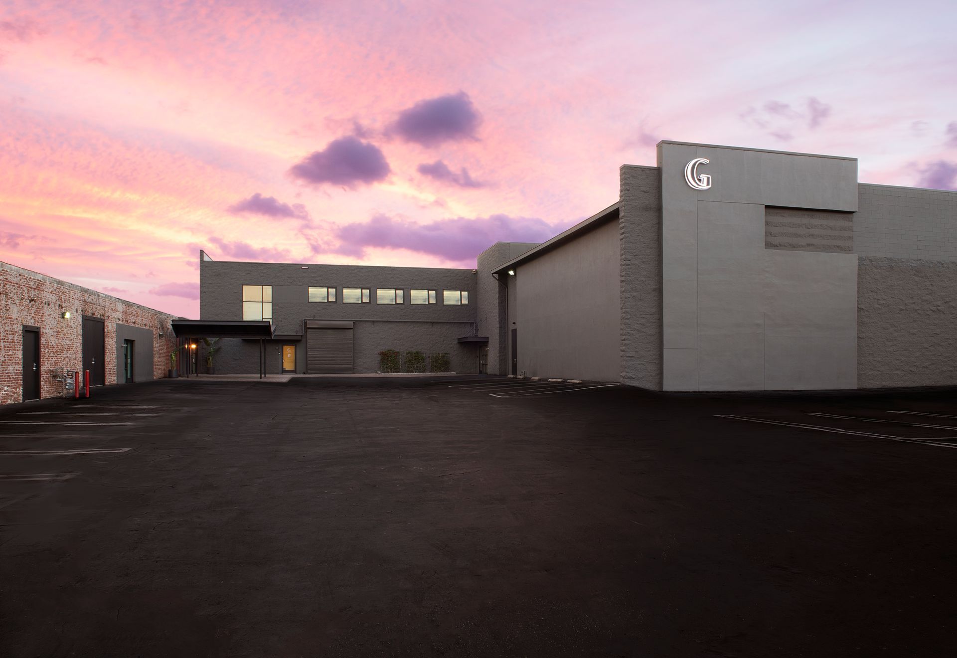 A grey industrial building with a large paved parking lot under a pink and purple sunset sky.