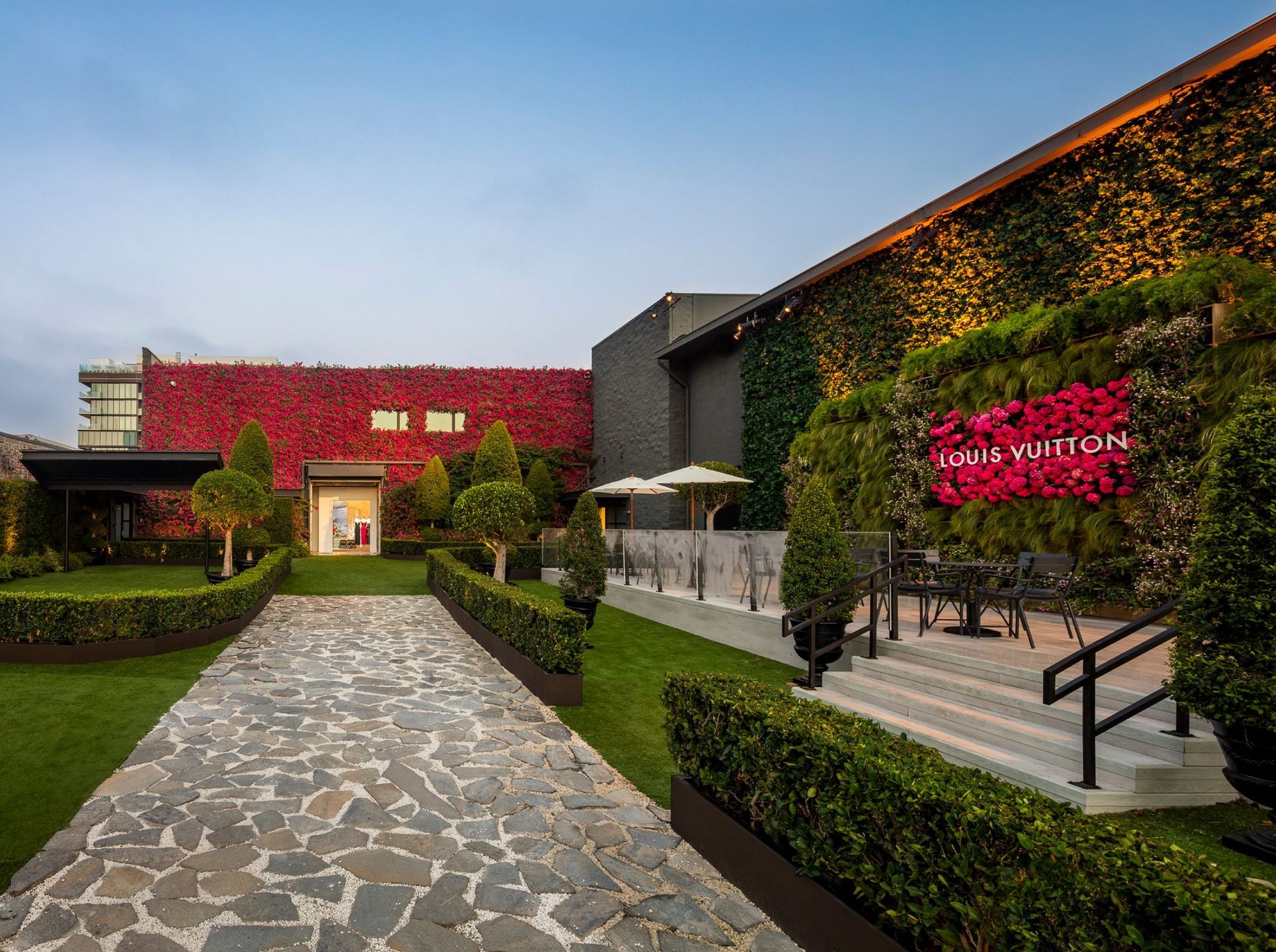 A stone walkway leads toward a Louis Vuitton building decorated with vibrant red flowering vines and green hedges.