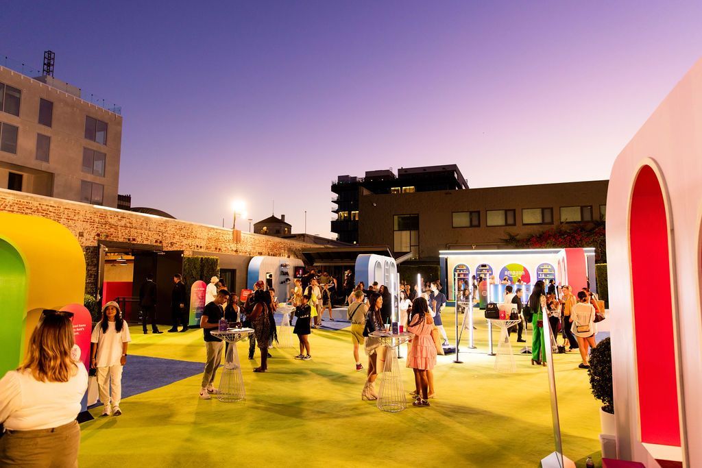 People gather at an outdoor evening event featuring colorful geometric arches, event displays, and a lime-green floor.