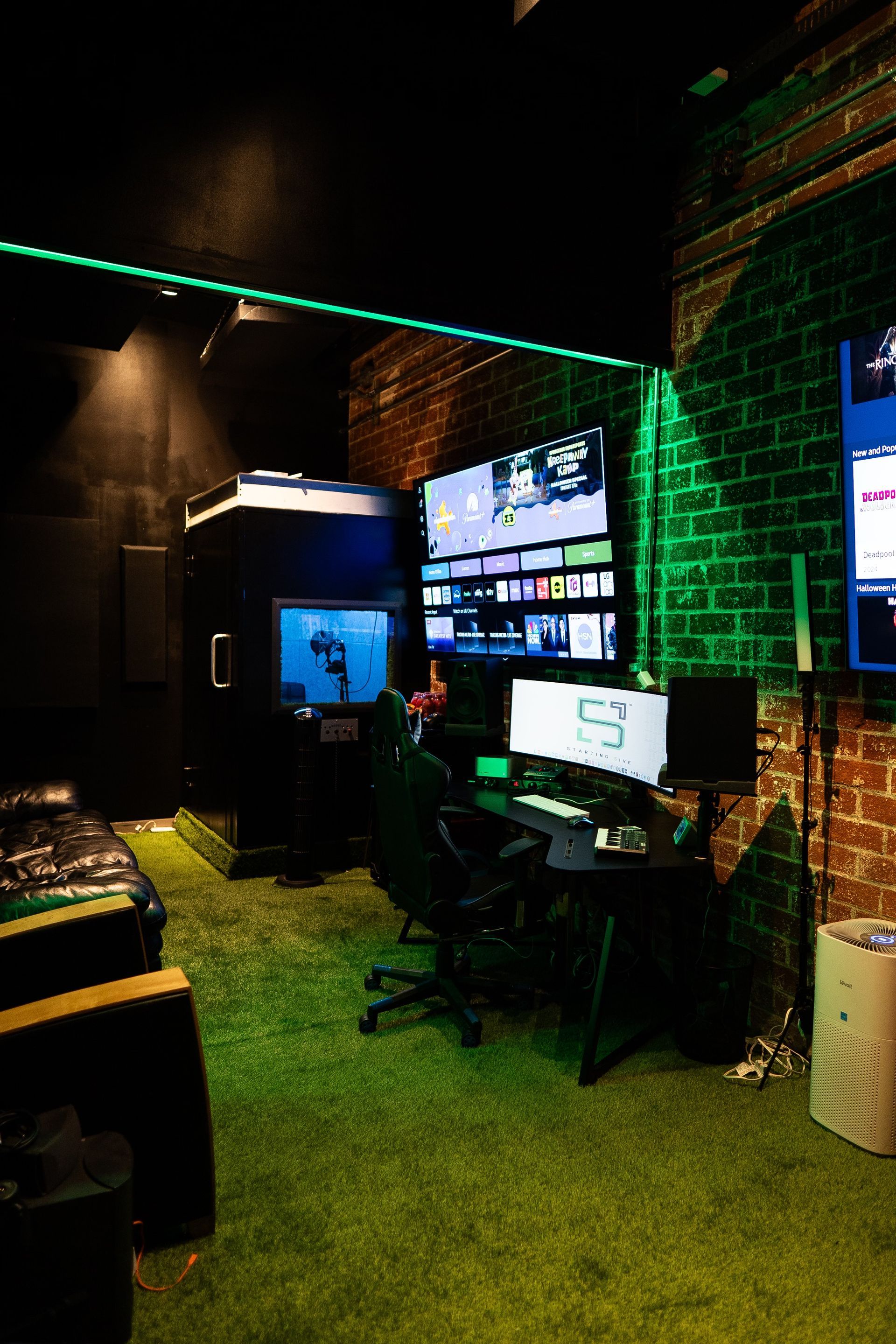 A gaming setup with a desk, multiple monitors, and a chair in a room with green artificial turf and exposed brick walls.