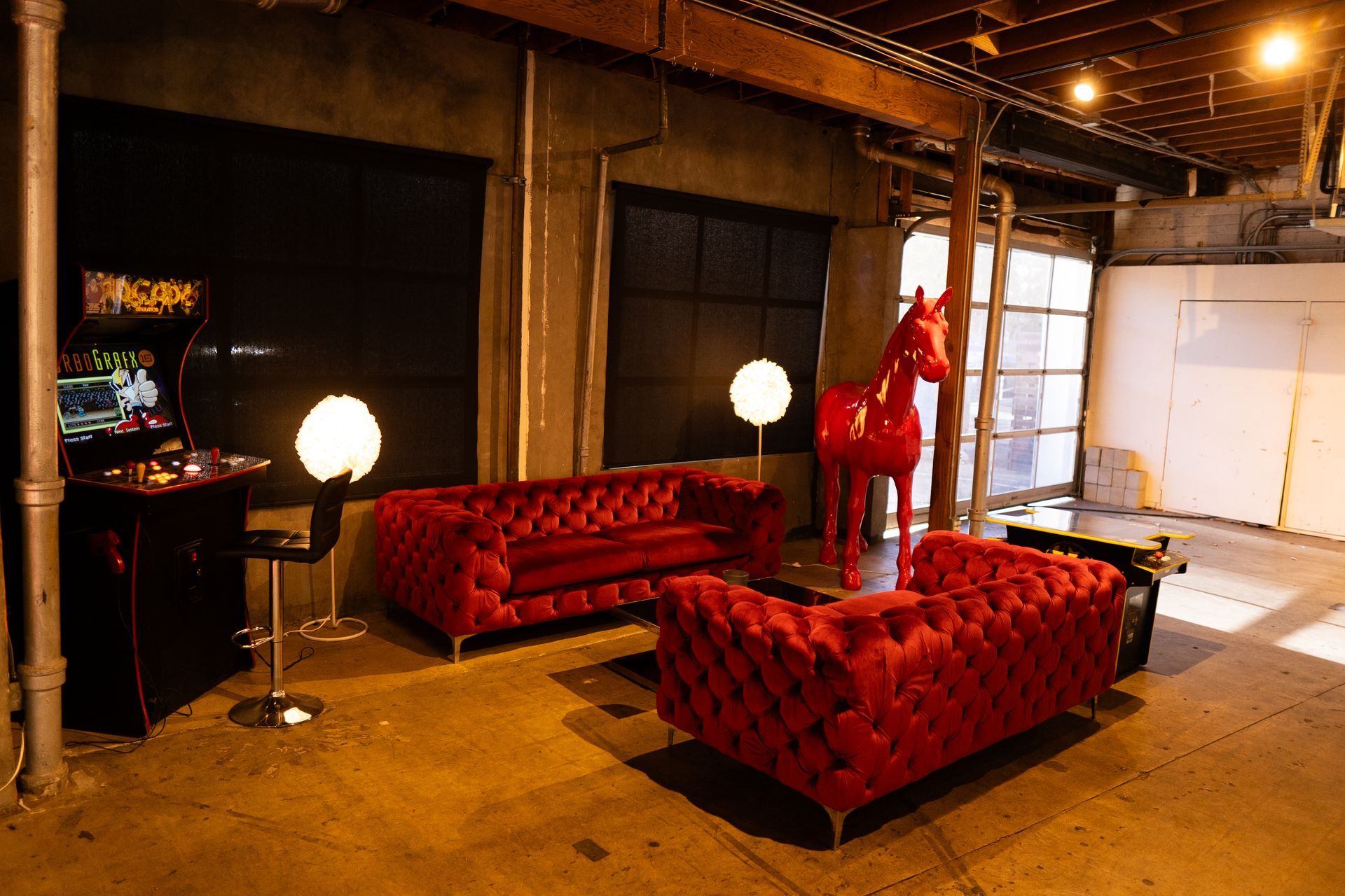 An industrial loft features two red velvet chesterfield sofas, a red horse statue, a retro arcade game, and globe lamps.