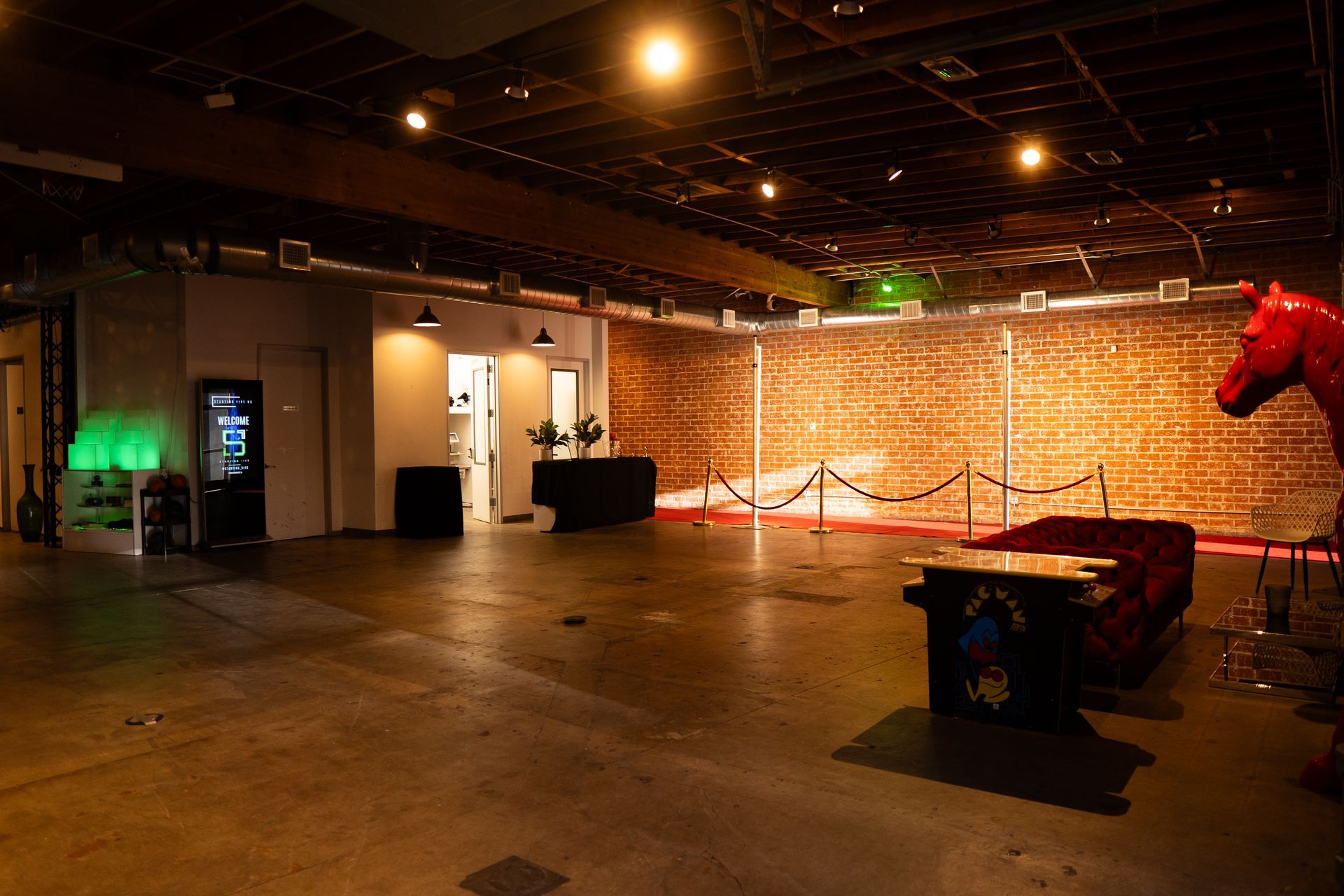 A dimly lit event space featuring a brick wall with string lights, a red horse statue, and a velvet sofa on a concrete floor.