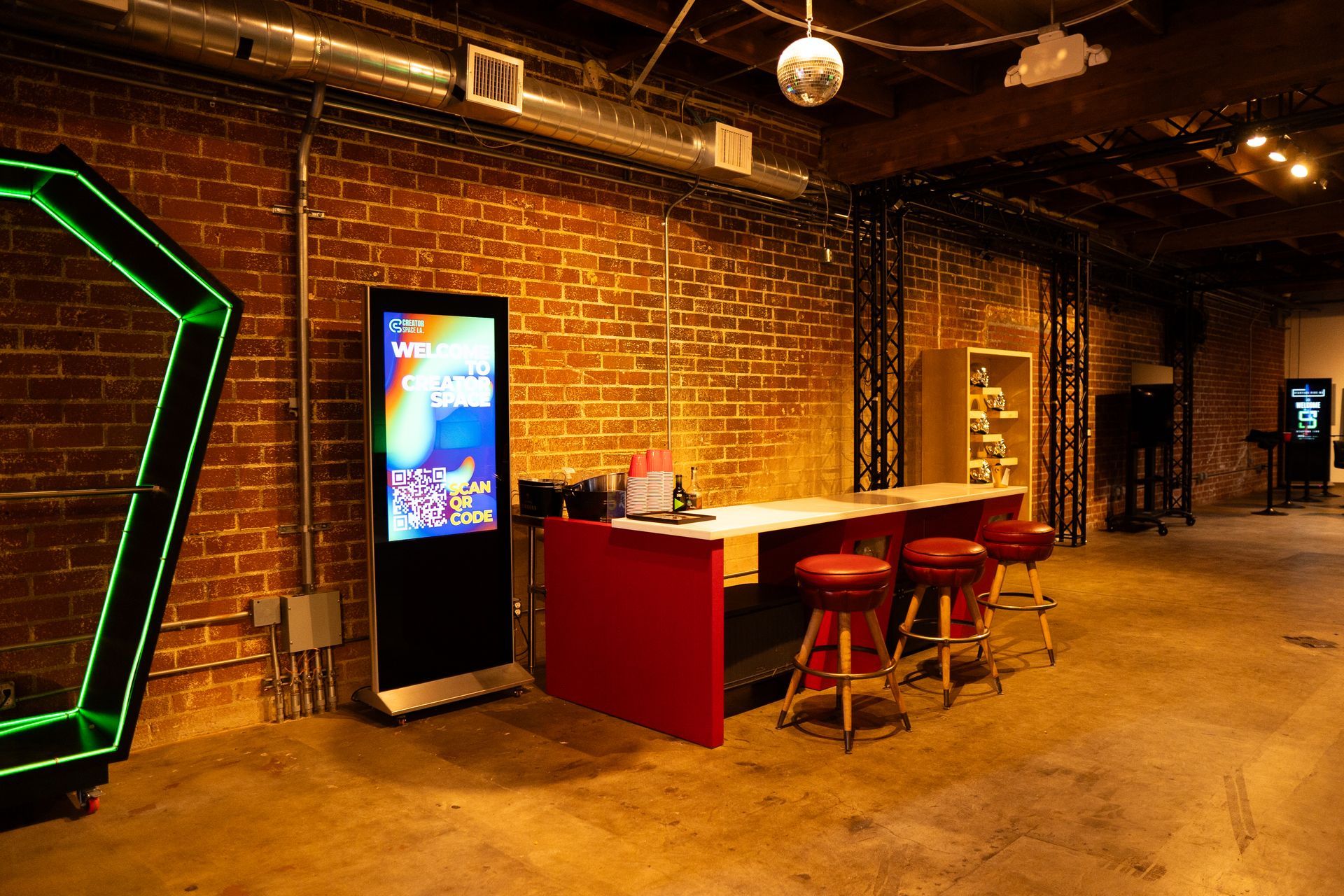 Red bar with three stools in a room with a brick wall, digital display, and a geometric green neon structure.