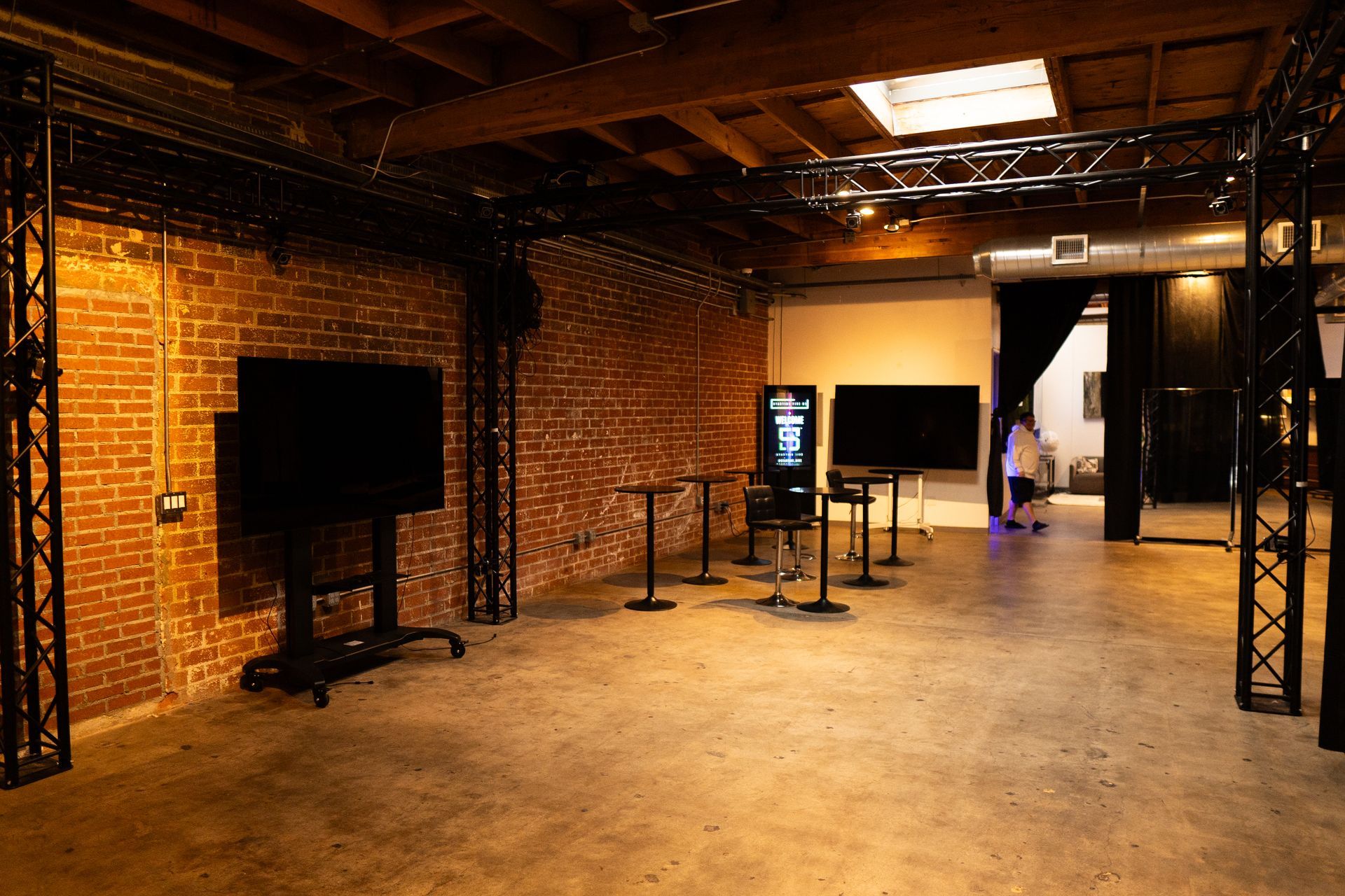 An industrial event space with brick walls, exposed wooden ceiling beams, metal trusses, floor TVs, and standing tables.