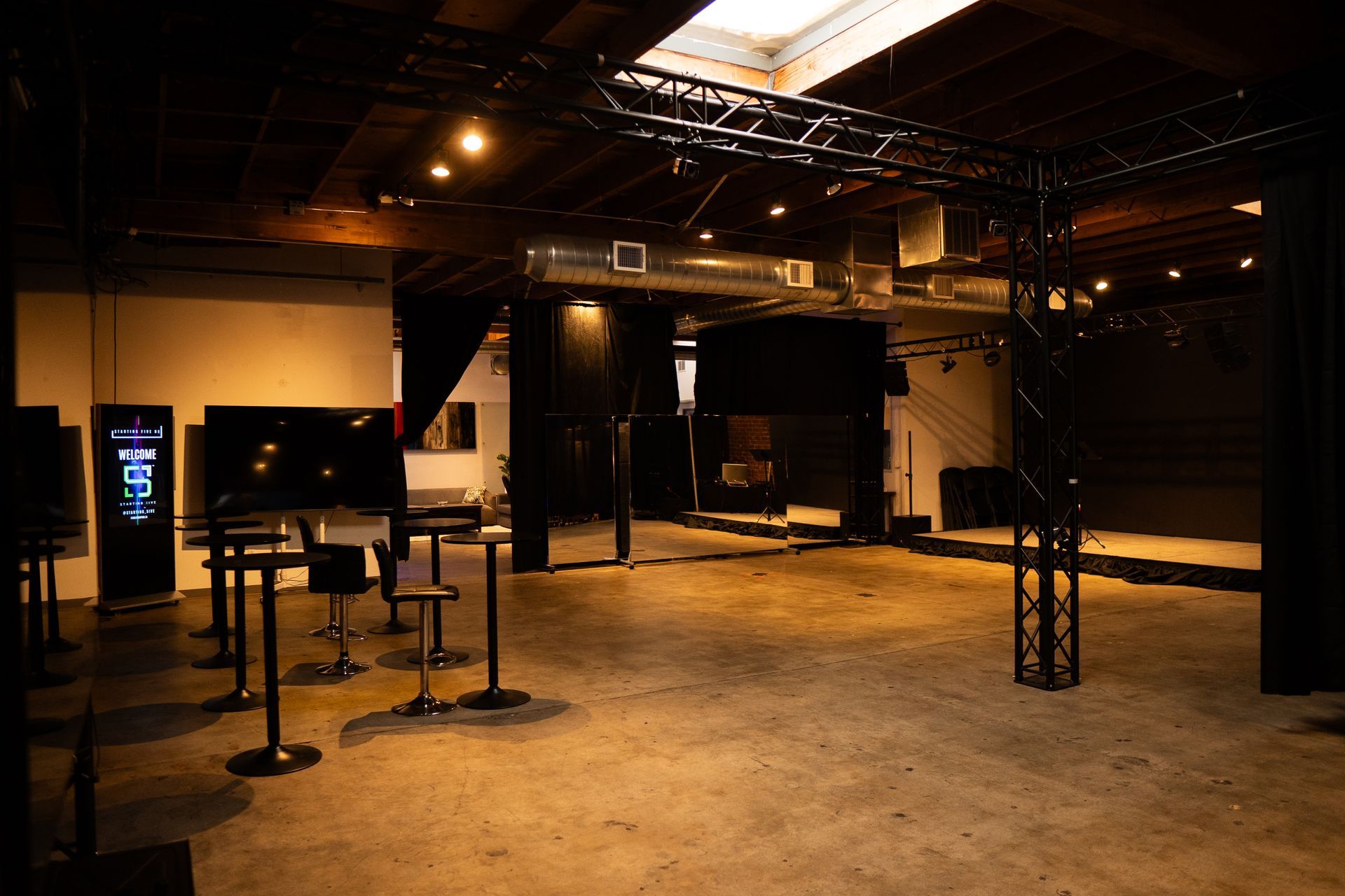 An industrial-style event venue with concrete floors, black metal trusses, high-top tables, and a stage area.